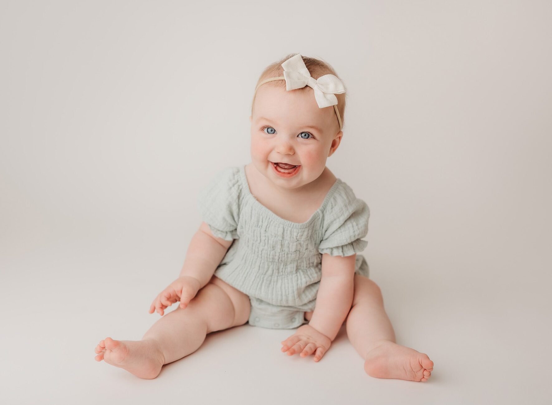 baby girl in teal dress sits on white backdrop for a sitter session in Nashville baby studio