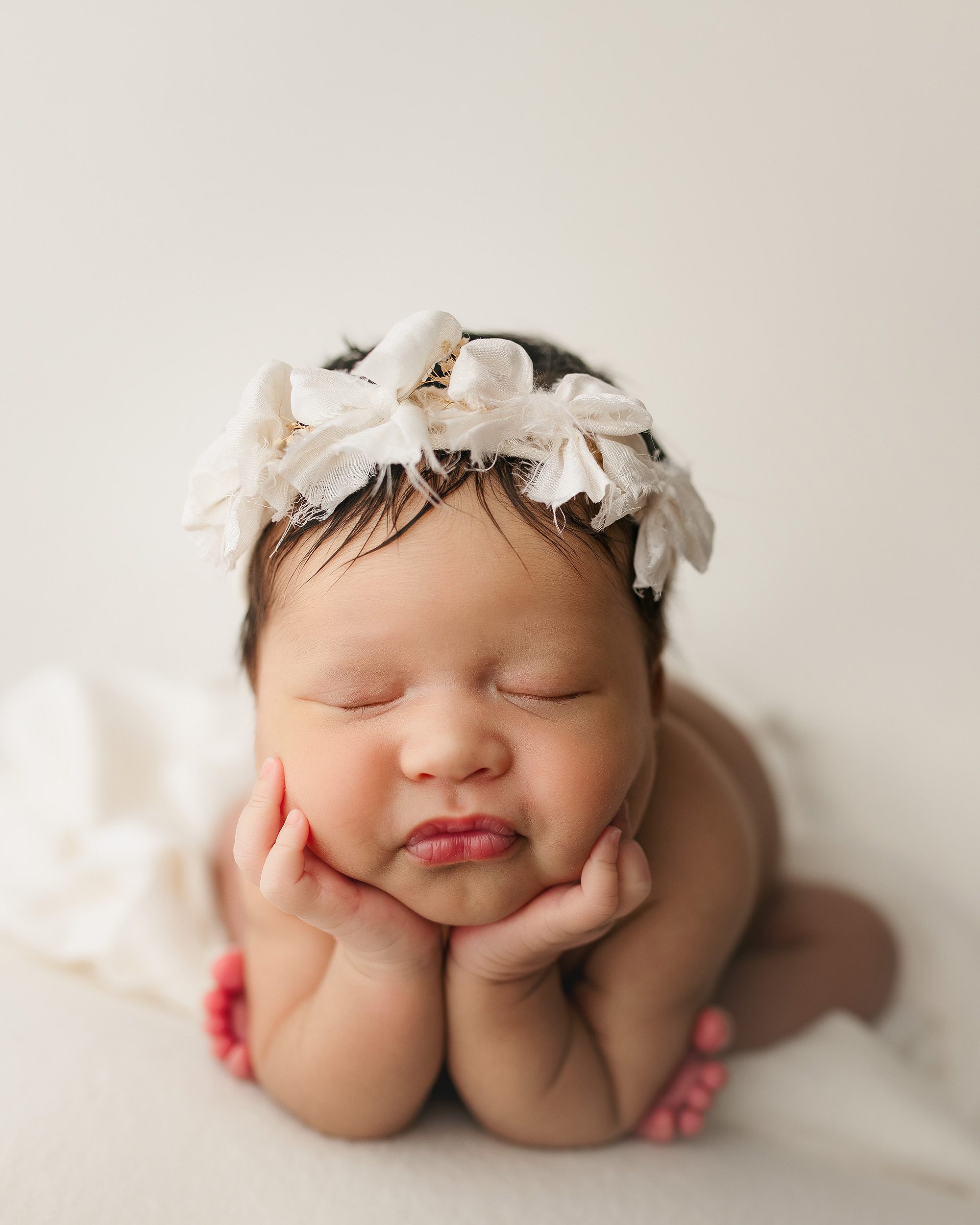 Newborn baby girl with white triple bow headband posed in froggy pose with hands under chin and legs tucked forward, sleeping peacefully on a cream blanket in a Nashville photography studio